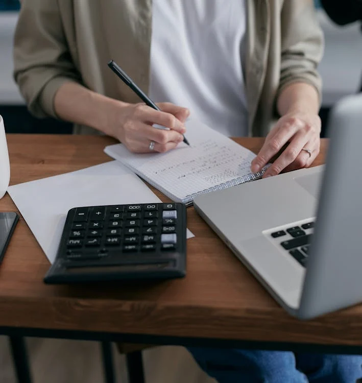 A woman sitting at a desk writing on a piece of paper with calculator and laptop.